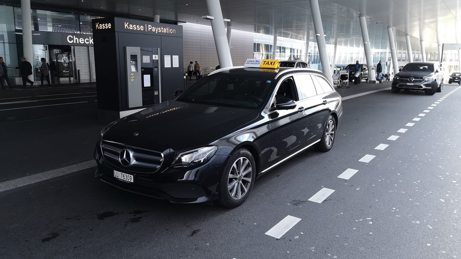 Schwarze Mercedes E-Klasse von Luzerner Taxi an der Vorfahrt eines Flughafenterminals