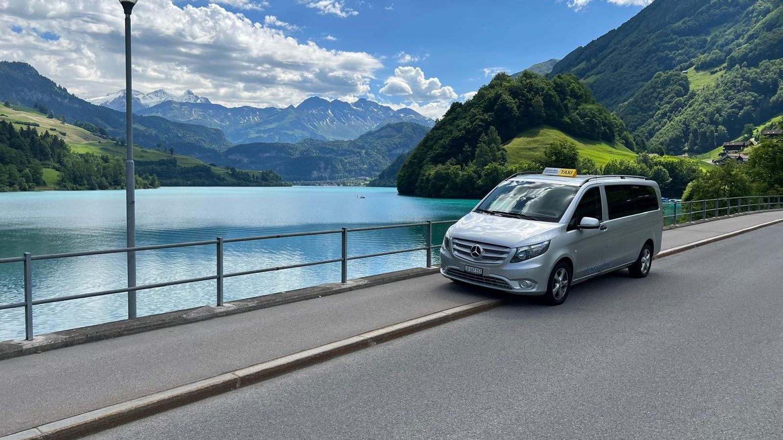Silberner Kleinbus von Luzerner Taxi auf einer Seestrasse unter den Bergen